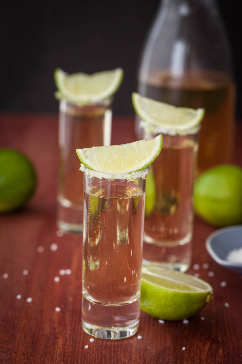 Three shots of tequila with lime wedges on a wooden table.