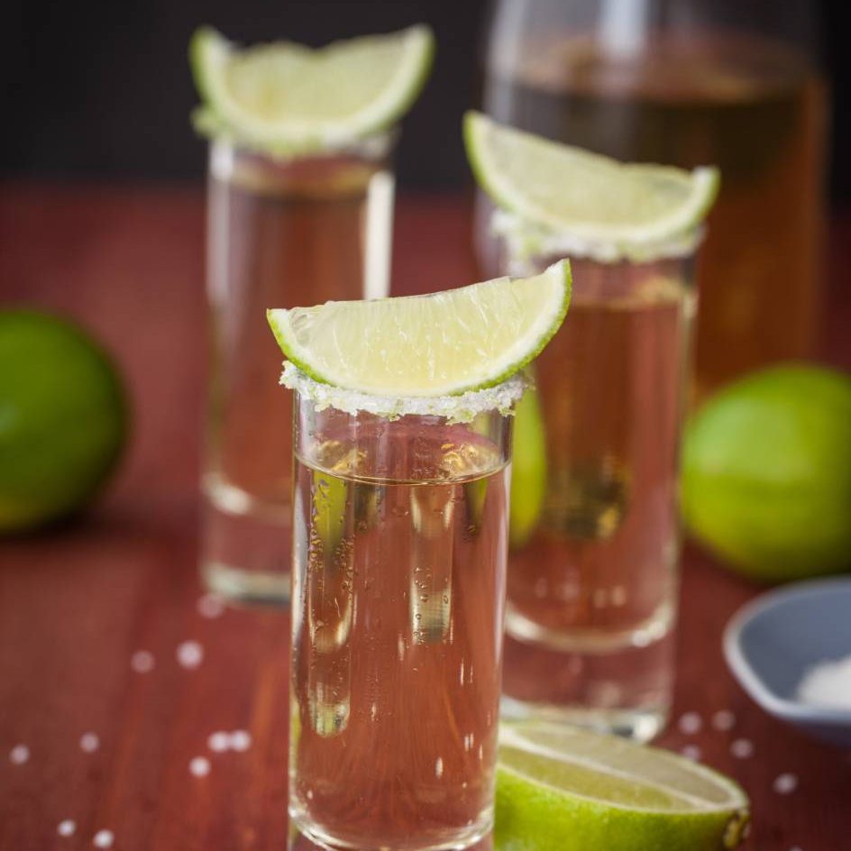 Three shots of tequila with lime wedges on a wooden table.